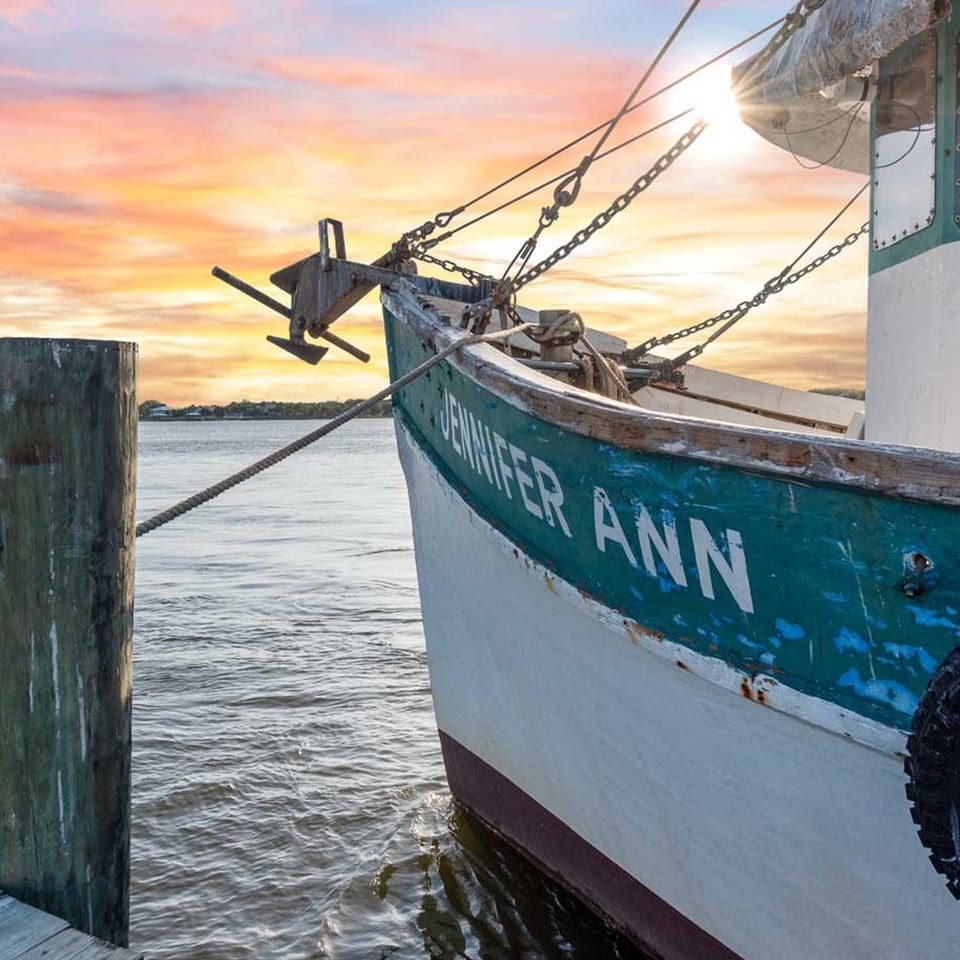 A fishing boat named "Jennifer Ann" docked at sunset with a vibrant orange and pink sky.