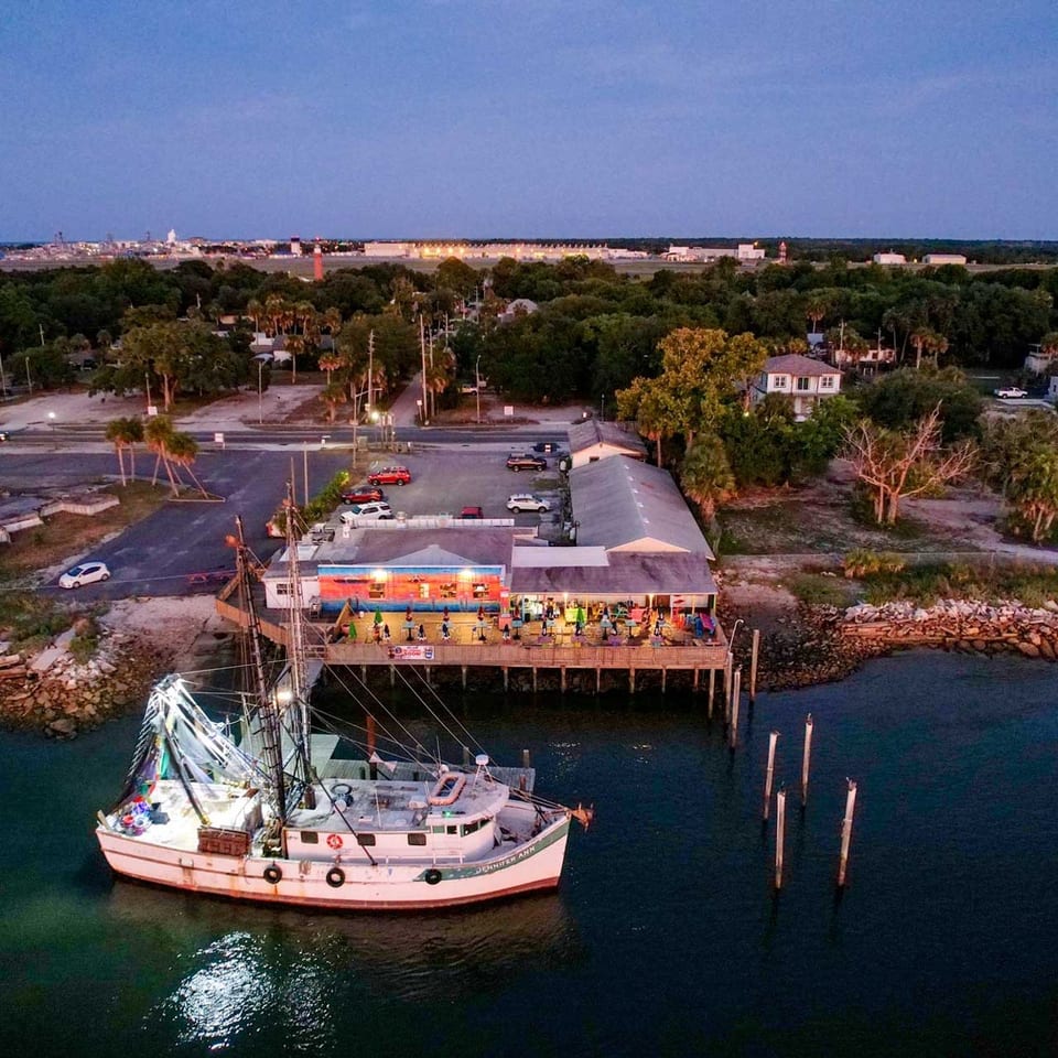 Aerial view of a boat docked next to a vibrant waterfront restaurant at dusk.