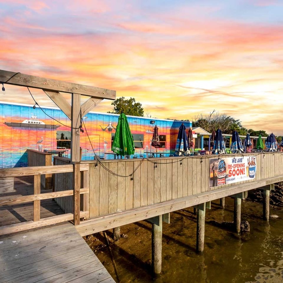 A vibrant waterfront restaurant with a colorful mural, outdoor seating, and umbrellas under a sunset sky.