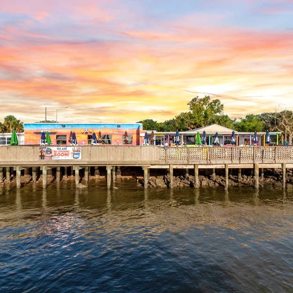 Waterfront restaurant with a colorful mural, outdoor seating, and a wooden deck under a vibrant sunset.
