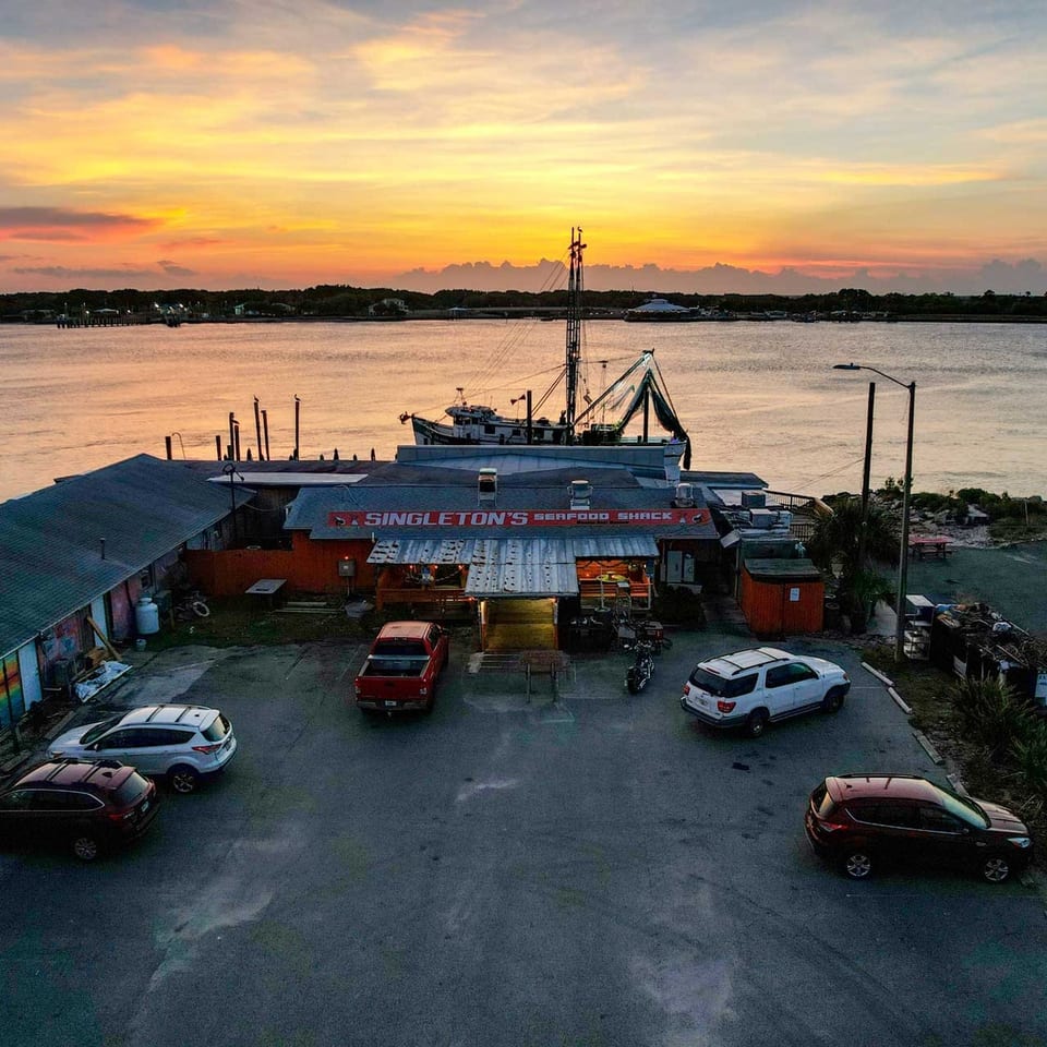 Singleton's Seafood Shack at sunset, with a fishing boat docked and cars in the parking lot.