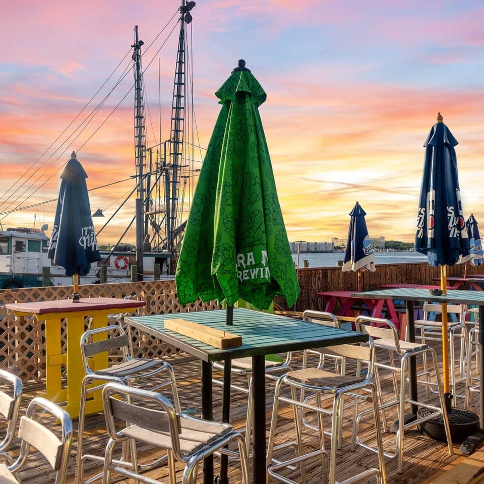 Outdoor restaurant patio with tables, chairs, and umbrellas overlooking a harbor with boats at sunset.