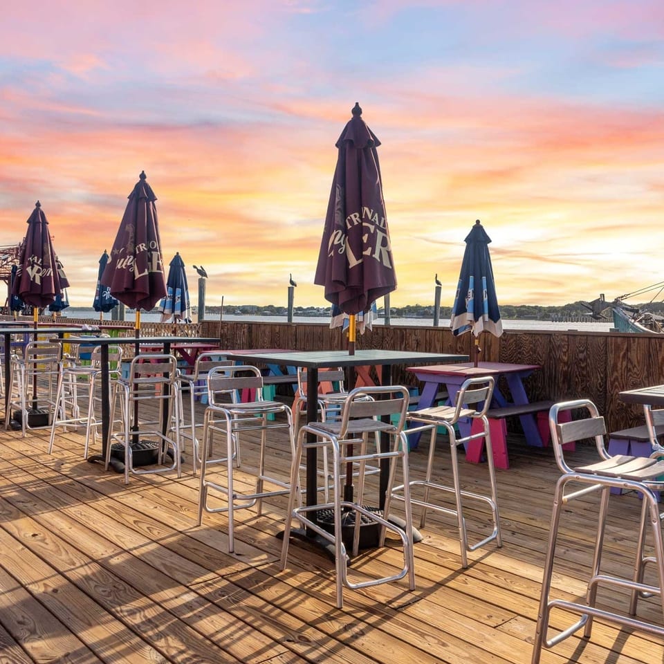 Outdoor restaurant patio with tables, bar stools, and umbrellas under a vibrant sunset sky.