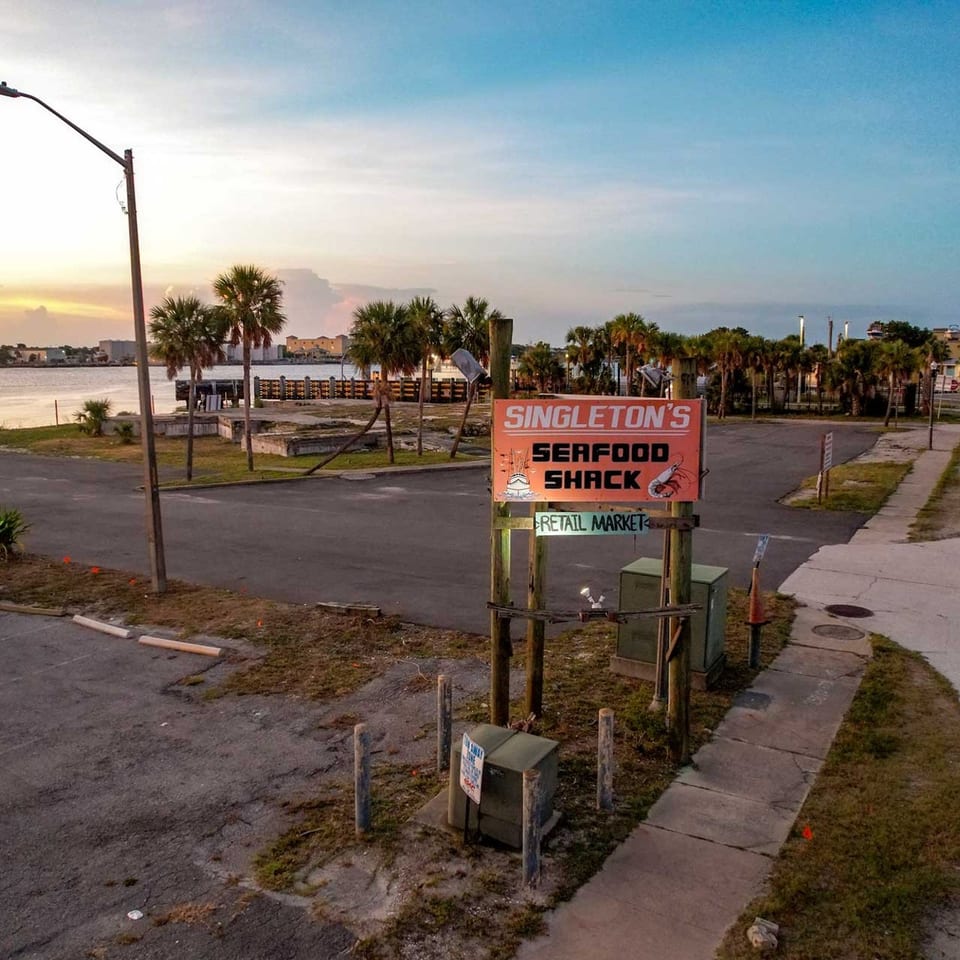 Singleton's Seafood Shack sign stands by a waterfront road with palm trees at sunset.