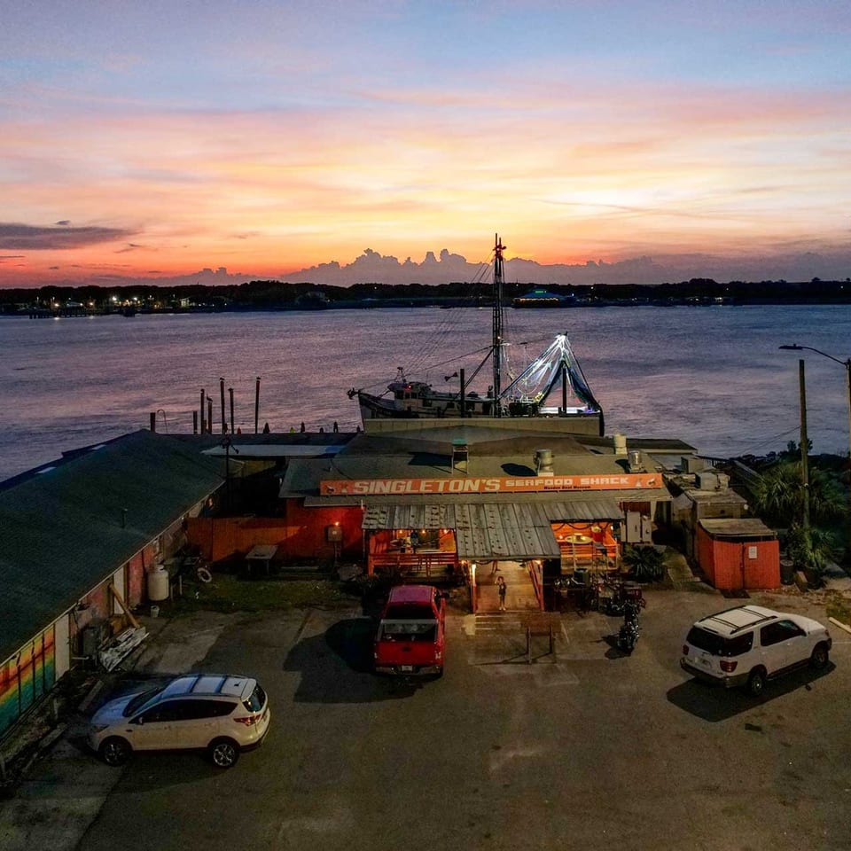 Singleton's Seafood Shack at sunset, with a fishing boat docked nearby and cars in the parking lot.