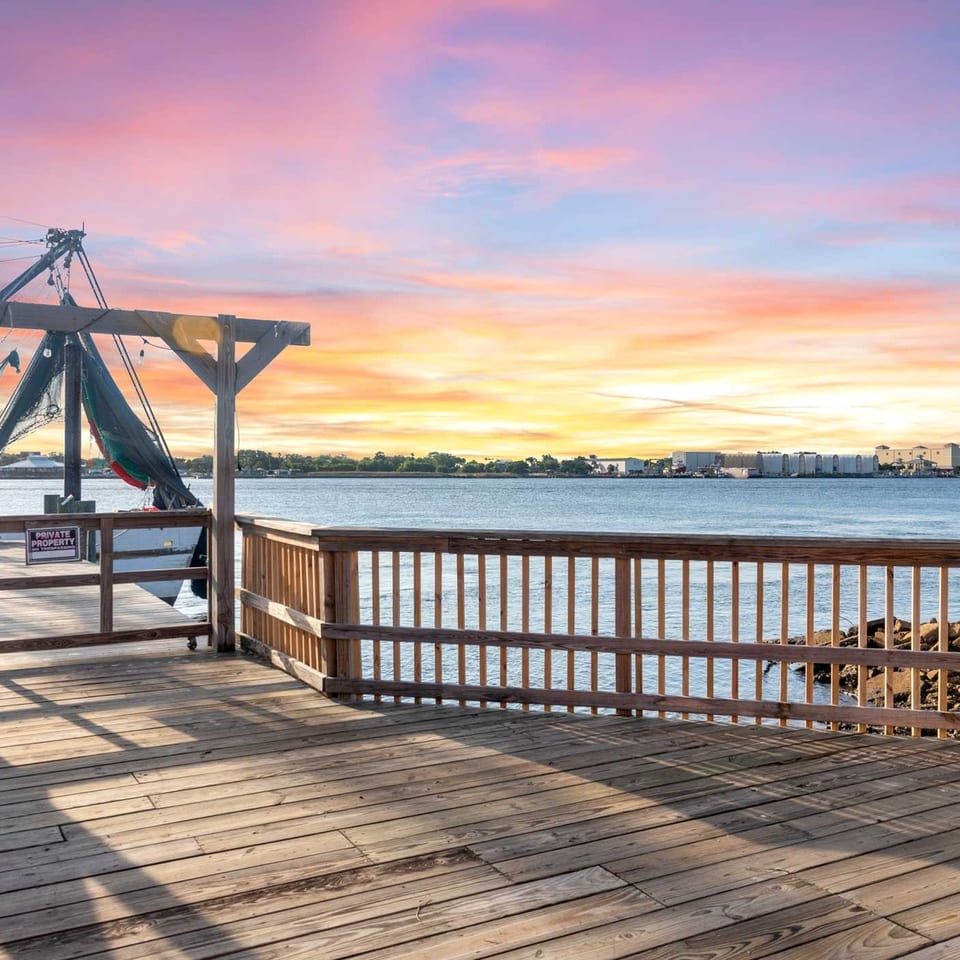 Wooden deck and railing overlooking a body of water with a shrimp boat and colorful sunset.