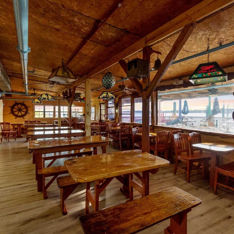 Rustic restaurant interior with wooden tables and benches, nautical decor, and large windows overlooking water.