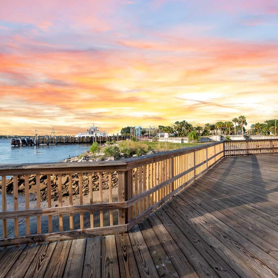 Wooden deck overlooking a harbor with a ferry, palm trees, and a vibrant sunset sky.